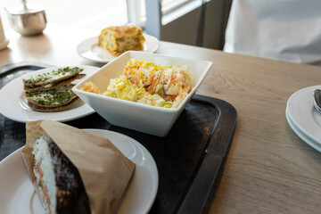 A close-up view of a lunch tray featuring a bowl of creamy coleslaw, a stack of savory sandwiches wrapped in brown paper, and a plate of baked goods.
