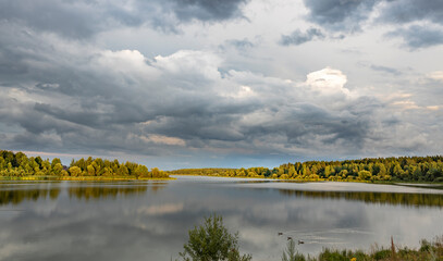 A picturesque scene of a serene lake nestled amidst a forested landscape, with a dramatic sky featuring a mix of dark, stormy clouds and soft, luminous patches of light.