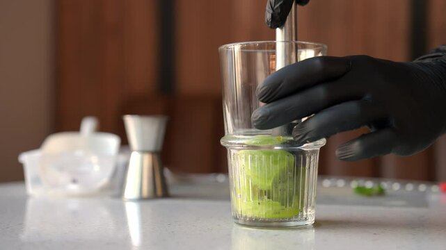 Professional bartender male in black gloves mashed with muddler kiwi and lemon slices in a glass to make a deliciously refreshing cocktail. Concept of party time, holidays, celebration mood.