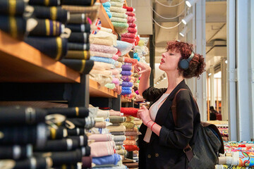 A woman is shopping for fabric in a store. She is wearing headphones and looking at the different colors and patterns of fabric