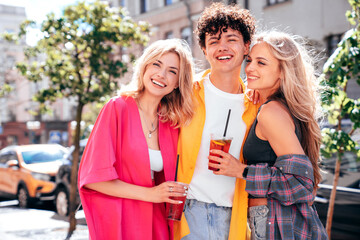 Young three stylish friends posing in the street. Fashion man and two cute female dressed in casual summer clothes. Smiling models having fun. Cheerful women and guy outdoors, Hold and drink lemonade