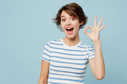 Young happy surprised shocked woman she wears striped t-shirt casual clothes showing okay ok gesture look camera isolated on plain pastel light blue cyan background studio portrait. Lifestyle concept.