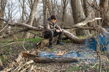 A teenage boy makes a bonfire in the forest, while hiking and outdoor activities, early spring landscape