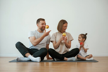 Obraz premium With apples in hands. Family of mother, father and daughter are doing yoga at home