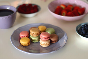 Plate of pastel macarons, cookies and chocolate, cup of tea of coffee, glass of bubble water, various berries, books and accessories on the table. Selective focus, pastel colors.