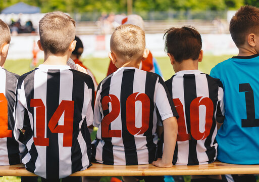 Soccer competition game for school kids. Children in sports team coached by young coach. Kids sitting on wooden bench during the half-time break. Soccer boys in sporty shirts with red numbers on backs