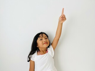 The little Asian girl raises her hand, points up with her index finger and smiles. A child in white...
