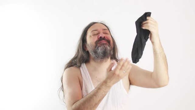 The poor man sniffs the sock and likes the smell. Satisfied facial expression. A man in a cheap, white, wrinkled undershirt. Bankrupt man without a job. 