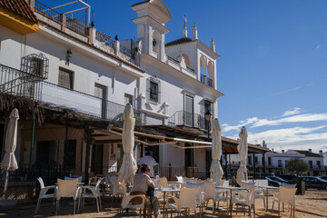 a sunny outdoor cafe scene in a charming European square, with patrons enjoying the ambiance under a clear blue sky, flanked by a classic white building with elegant architectural details.