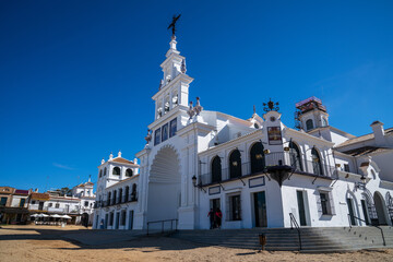Obraz premium The pristine facade of the Hermitage of El Rocío gleams under the Spanish sun, with its elegant white walls and ornate religious detailing standing out against the clear blue sky.