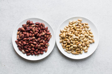 Overhead view of unpeeled raw peanuts and peeled roasted peanuts on white plates, flatlay of raw and roasted peanuts on a marble countertop, process of making roasted peanuts