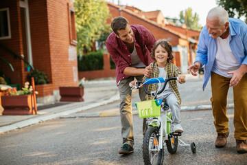 Father and grandfather teaching young boy to ride a bike on residential street