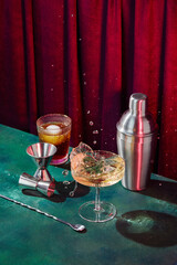Splash in champagne glass, cocktail glass, shaker, cocktail mixing spoon and jigger on dark green table in disco club atmosphere