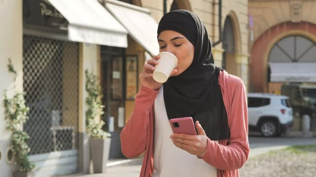 arab female using smartphone walking down city street slow motion,middle easter girl wearing hijab walks in the town uses smart phone and drink coffee