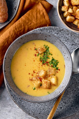 Bowl of mashed potato soup with a spoon on the plate. The bowl is filled with yellow soup puree and topped with croutons, parsley and ground red pepper.