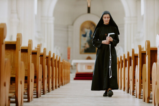 Wide shot of senior Caucasian nun holding Bible book walking along nave in Catholic church, copy space