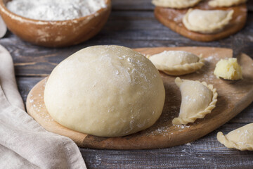 Dough for dumplings on a wooden cutting board, ready-made dumplings, a bowl of flour and a spoon with potato filling on a wooden table