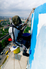 on the roof of a high-rise building against the background of multi-colored bright houses painter suspended on a safety net paints the wall blue