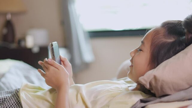 Cheerful Asian child using smartphone in cozy bedroom, highlighting the child's relaxed posture as lie comfortably in bed. Their face displays pure enjoyment, comfort for connection modern technology.