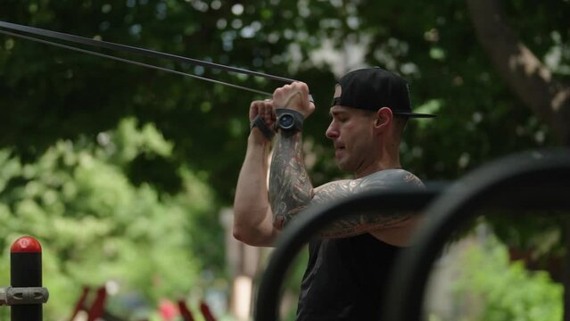 A fit, tattooed man is working out in an outdoor gym park setting doing tricep extensions. He is wearing a black sleeveless shirt, black baseball cap and a watch