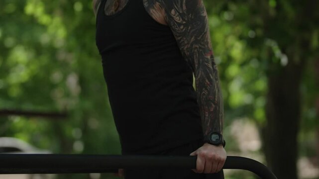 Fit, tattooed man doing dips on parallel bars in a park. He is working out on a sunny day, wearing a fitness tracker.