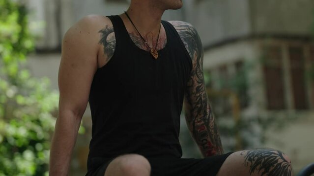 A young, fit, tattooed man with a black hat on backwards and wearing a necklace sits and talks, waiting for someone outdoors on a summer day.