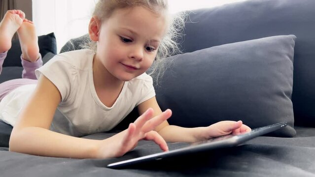 Little girl using tablet device, lying on sofa. Smiling little girl watching online cartoons on tablet on Internet using digital tablet technology device. 