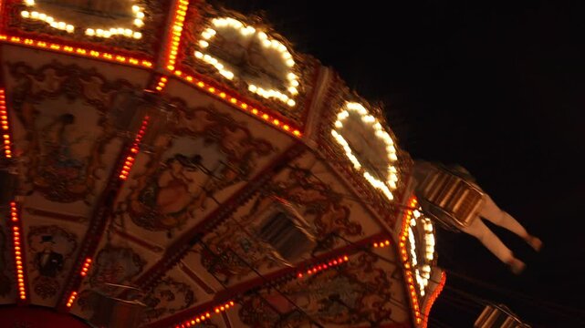close-up night view of an ornate carousel in motion, illuminated by bright lights against a dark sky. The carousel's intricate designs and vibrant colors are highlighted by rows of lights that create