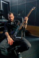 Fototapeta premium in the equipment of a recording room handsome man with a beard and tattoos sitting posing for the camera with a guitar