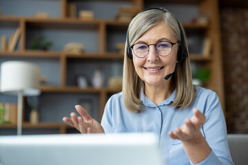 Smiling senior woman in a home office setting, wearing a headset, and gesturing while video...