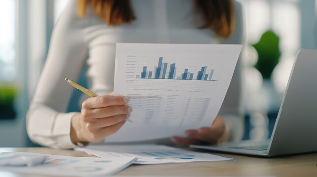 Businesswoman analyzing financial report at office desk, reviewing charts and graphs for company performance on laptop.