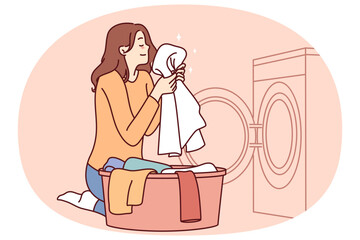 Housewife woman sits near washing machine and inhales fragrant smell freshly washed towel after using good laundry detergent. Girl takes out washed clothes from washing machine putting them in bowl