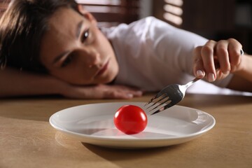 Eating disorder. Sad woman holding fork with tomato at wooden table indoors, selective focus