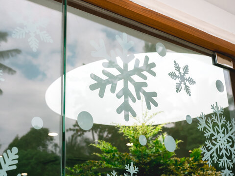 Christmas snowflake sticker decoration on glass window in cafe, reflection with green tree, outdoor garden and blue sky background.