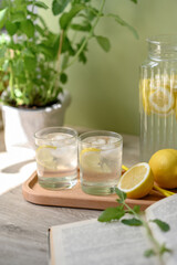 Glasses with summer lemonade drink, lemons, jug, open book, mint plant on table with natural sunlight, neutral beige and green background
