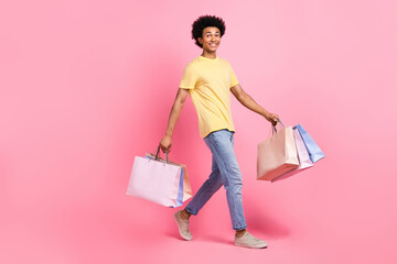 Full length photo of pretty excited man wear yellow t-shirt walking holding bargains empty space isolated pink color background