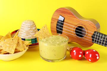 Delicious guacamole with nachos chips, Mexican sombrero hat, ukulele and maracas on yellow background