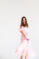 Model wearing casual outfit. Look in trendy minimalistic style. Pink dress with slit, belt, puff sleeves. Sexy sensual happy woman. Positive woman turn around and hold pink flower on white background.