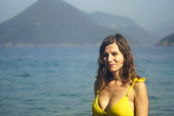 A smiling slender woman, dressed in a two-piece swimsuit, stands against the backdrop of the bright waters of the Adriatic Sea and a picturesque mountain. Beach holiday in Lustica, Montenegro.