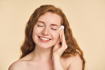 Woman enjoying her skincare routine, holding a cotton pad against her cheek. Radiant skin, self-care, and happiness concept on a warm beige background.