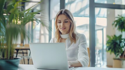 A woman working on her laptop in a modern office setting, with natural light and greenery in the background.