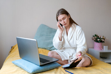 A woman in a robe is talking on her cell phone while sitting on a bed