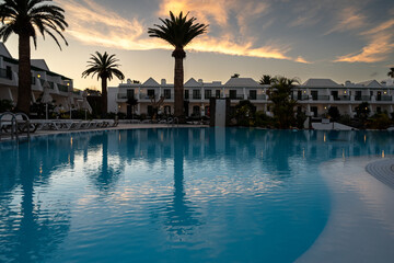 A pool with a palm tree in the background. The pool is surrounded by buildings. The sky is cloudy and the sun is setting