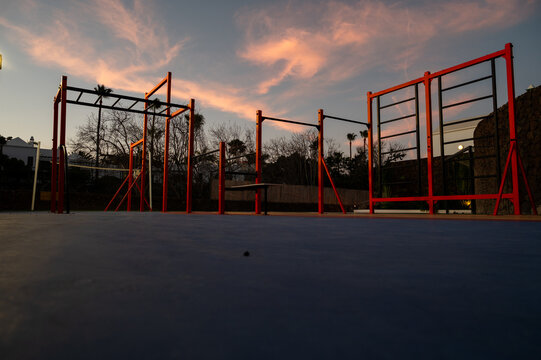 A park with a lot of equipment and a beautiful sunset in the background. The equipment includes a pull up bar, monkey bars, and a bench