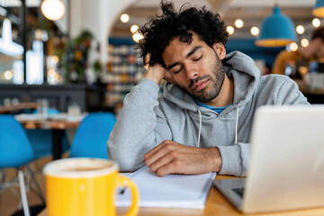 Fototapeta premium A man sleeping at a cafe table with a laptop in front of him. A cup of coffee is in the foreground, and the mans head is resting on his arm.