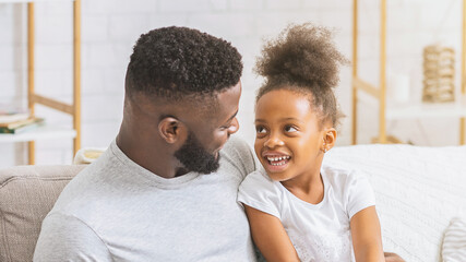 A close-up image of a happy Black father and daughter laughing together on a couch in a living room. The two are enjoying each others company and are clearly having a good time.