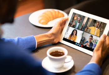 Black man sits at a table in a casual setting, participating in a video conference on a tablet. The screen displays four participants, each in a different video frame.