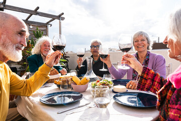Group of happy senior friends bonding at home for dinner party - Cheerful and youthful old mature multiethnic people having fun and dining on a rooftop terrace