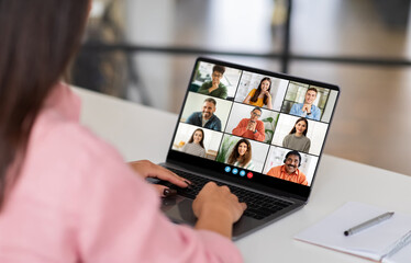 A woman sits at a desk in a home office, looking at her laptop screen as she participates in a group video conference. The screen displays several participants in a grid format, webinar