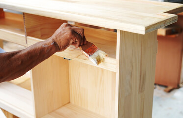 Carpenter hands applying varnish to a cabinet with a paintbrush. First lacquer coating on wood surface at a furniture and wooden workshop.Close up.